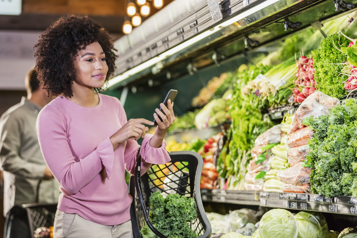 A young African-American woman in her 20s shopping in the produce aisle of a supermarket. She is using a mobile app on her smart phone for her shopping list.