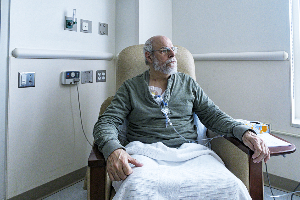 A recovering senior adult man colorectal cancer patient is sitting resting comfortably in a hospital cancer ward easy chair while chemotherapy IV drip medicine is administered by an array of medical equipment through a subcutaneous intravenous chemo access port temporarily embedded into his upper chest. "Daily Living With Cancer" image brief - #700034767.