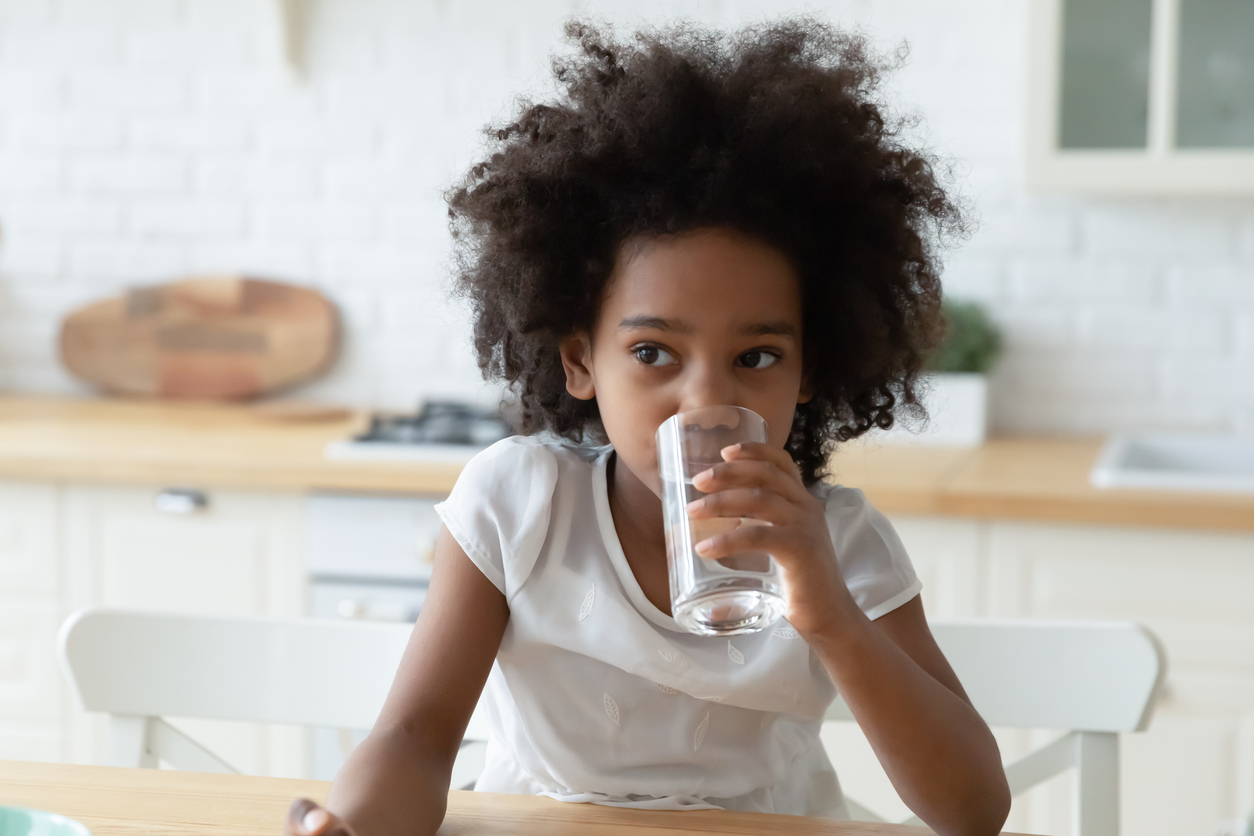 Thirsty small African American girl child sit at home kitchen feel dehydrated enjoy clean clear pure mineral water from glass. Little teen ethnic kid sip aqua for body refreshment. Hydration concept.