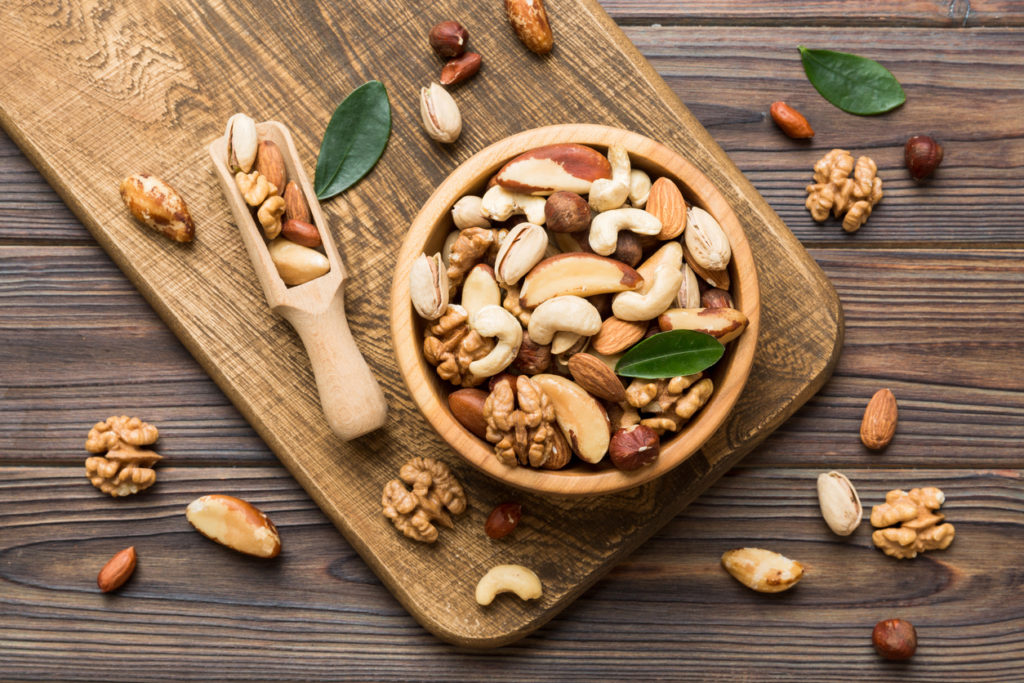 Mixed nuts in a wooden bool surrounded by additional nuts and a scoop